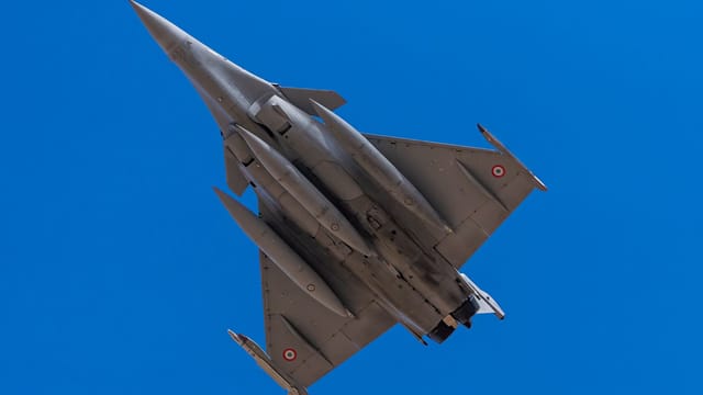 Dassault Rafale fighter jet from below against a bright blue sky.
