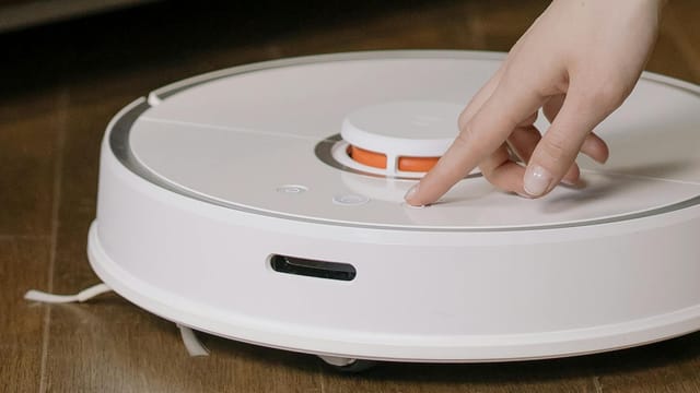 Close-up of a hand pressing a button on a robotic vacuum cleaner on a wooden floor.