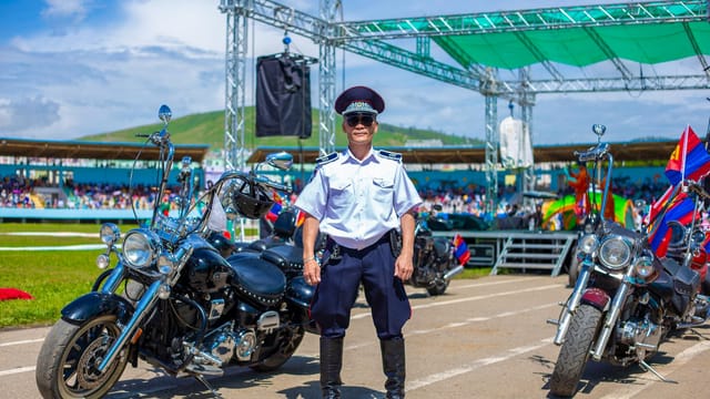 Police officer poses with motorcycles during an outdoor event with flags and a clear sky.