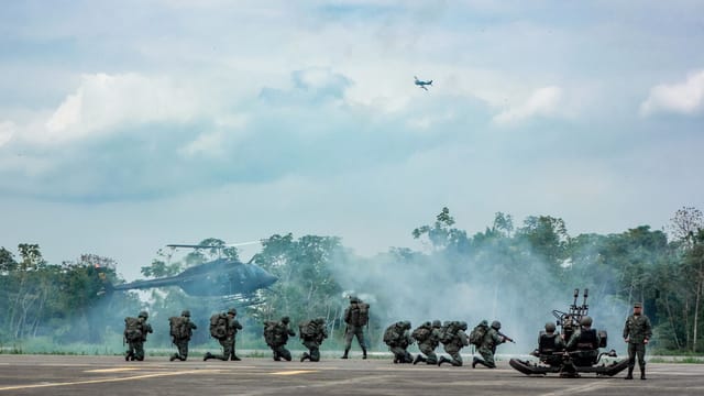 Back view of anonymous army of soldiers in uniforms with shotguns shooting while standing leaned on one knee on asphalt pavement near helicopter and trees under cloudy sky