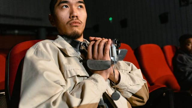 An adult male sitting in a cinema, holding a video camera, with red seats around.