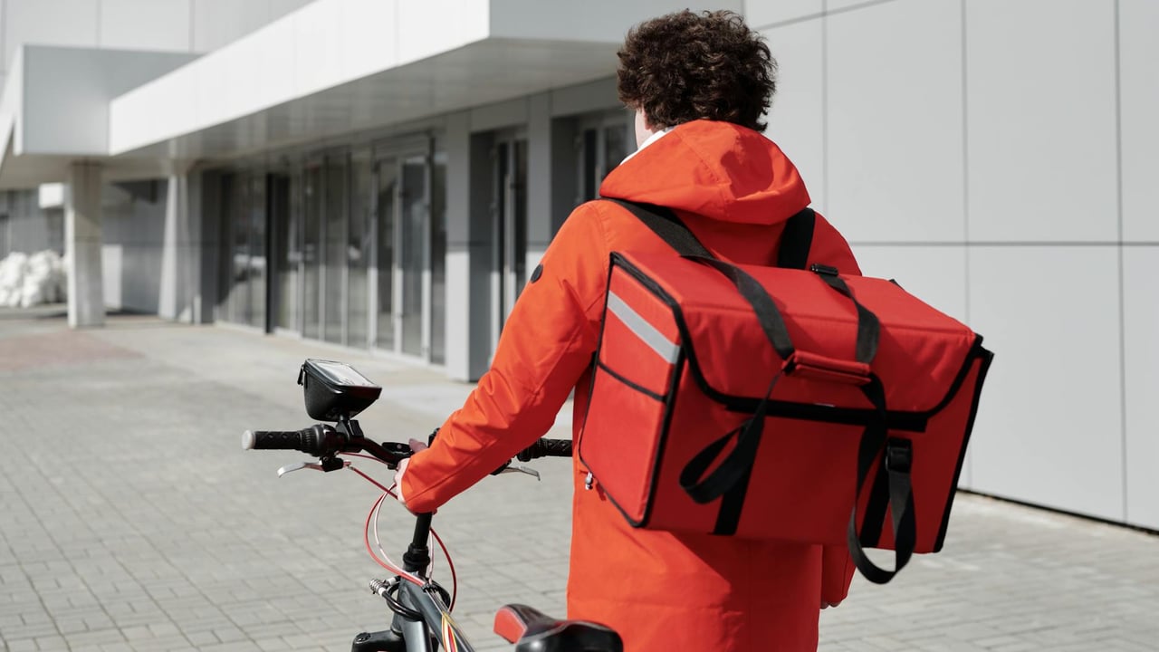 A food delivery rider in a red jacket walking a bike with a thermal bag outdoors.