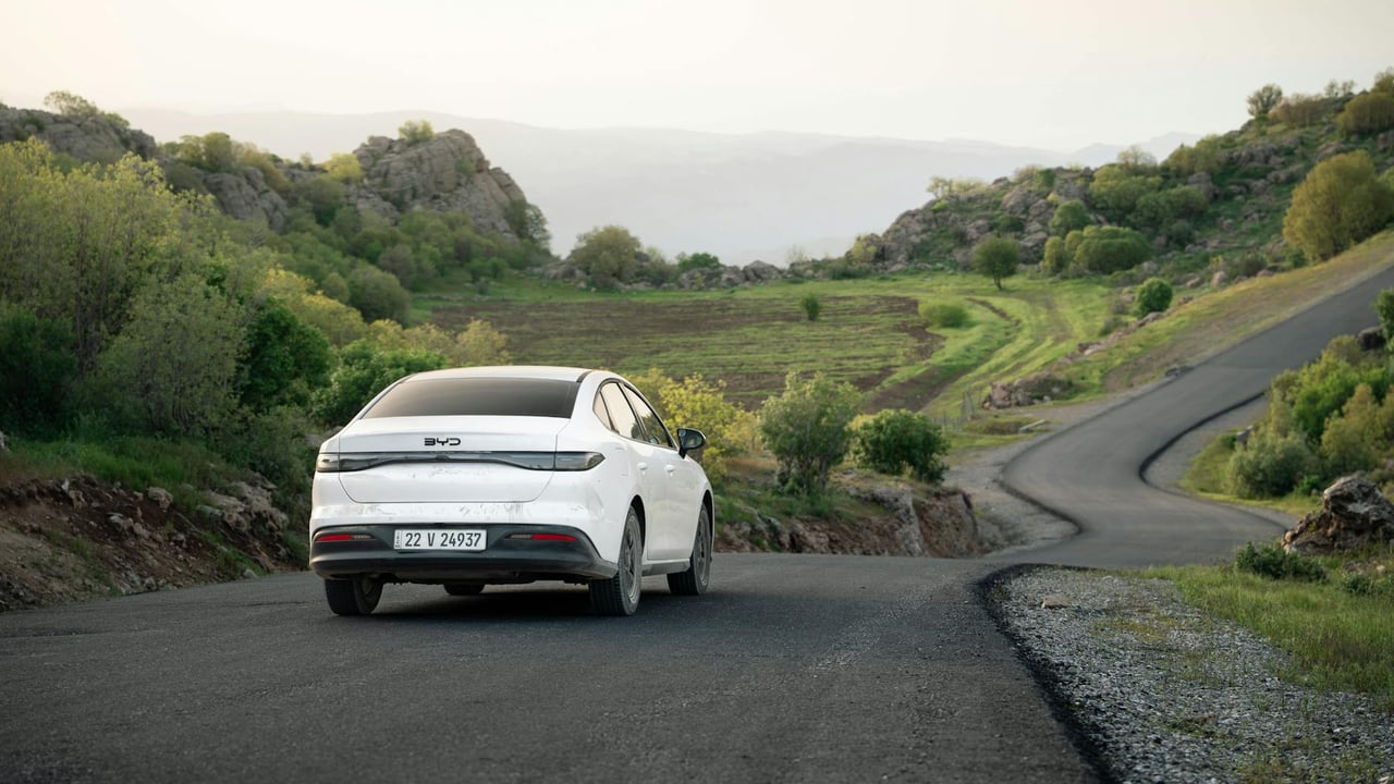 A white car drives on a winding road through scenic hills in Shaqlawa, Iraq during daylight.