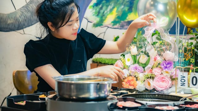 A young woman arranges flowers in a warm Vietnamese diner setting with a hotpot meal.