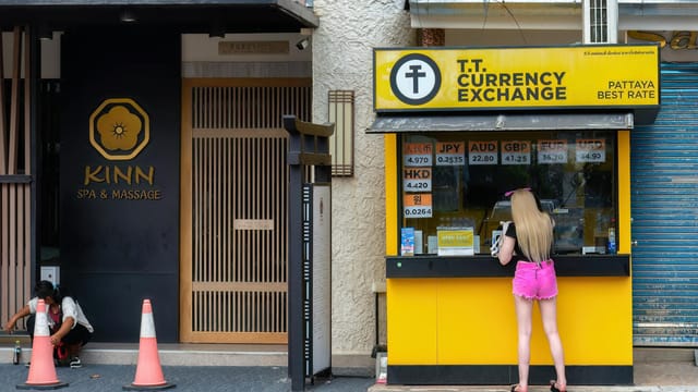 Woman at currency exchange booth in Pattaya, Thailand, with currency rates displayed.
