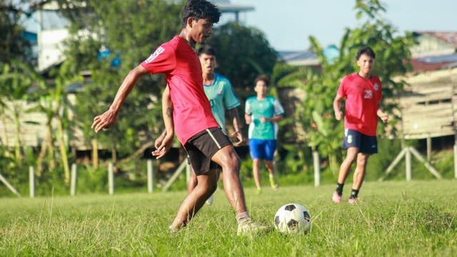 A group of young men enthusiastically playing soccer on a lush green field in Myawaddy, Myanmar.