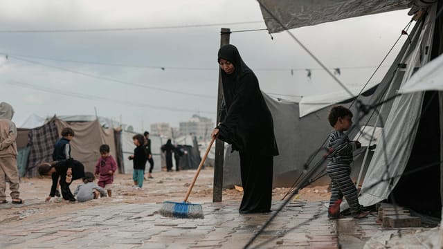 A woman sweeps in a Gaza refugee camp with children playing nearby.