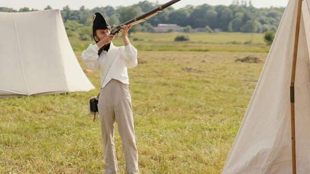 A soldier in historical attire aims a musket during a reenactment on a grassy battlefield.