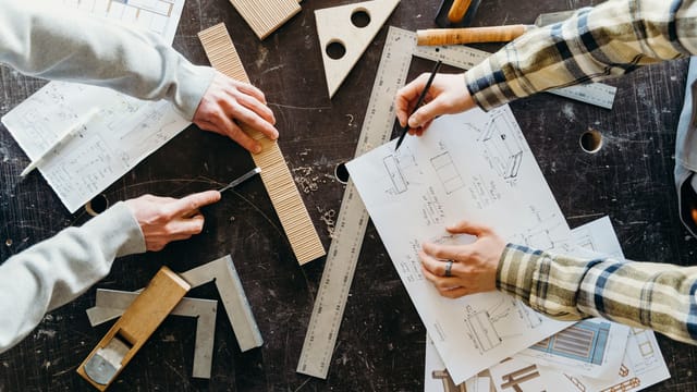 Top view of two people designing and crafting woodworking projects with blueprints and tools.