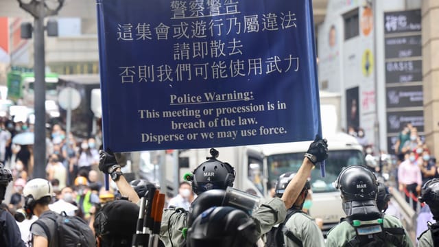 Policemen with shields holding warning sign during crowded protest.