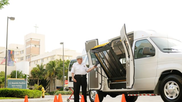 A security guard operates a wheelchair lift on a van outside a medical facility.