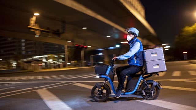 A masked delivery driver rides an electric scooter on Wuhan city streets at night.