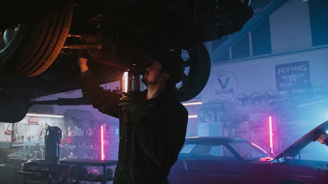 A mechanic inspects a car's undercarriage in a vibrant, neon-lit garage setting, highlighting automotive repair work.