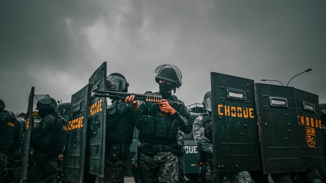 Riot police in tactical gear holding shields during a stormy protest.