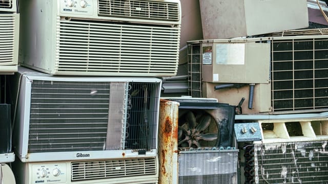 A collection of broken and rusted air conditioners piled in an outdoor junkyard.