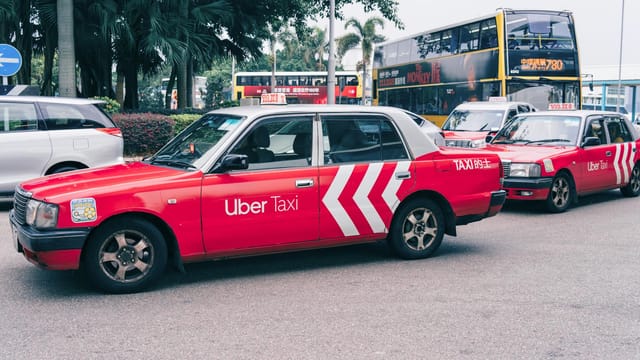 Red Uber taxi parked on a bustling city street with buses and greenery in the background.