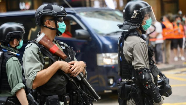 Police officers wearing riot gear and face masks stand in an urban street with protective equipment.