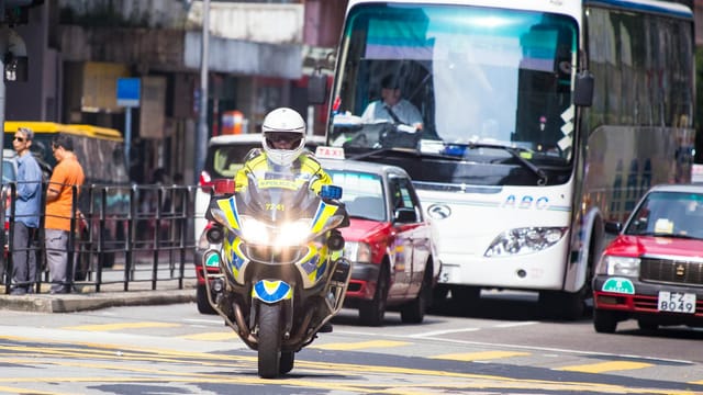 A Hong Kong police officer on a motorcycle navigating street traffic during the day.