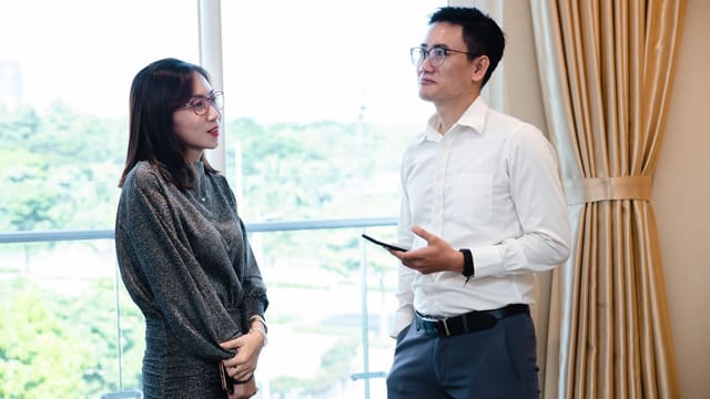 Two colleagues engaged in a business conversation indoors with a window view.