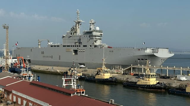 Aerial view of a large navy ship docked at Lisbon harbor alongside tugboats on a clear day.