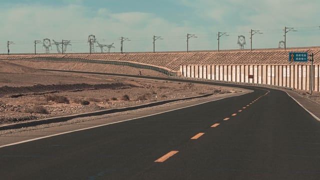 A lonely road curves through the desert with railway lines and signs in Chinese script.