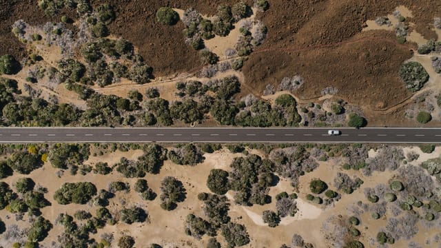 An aerial shot capturing a lone car driving on a straight desert road surrounded by sparse vegetation.
