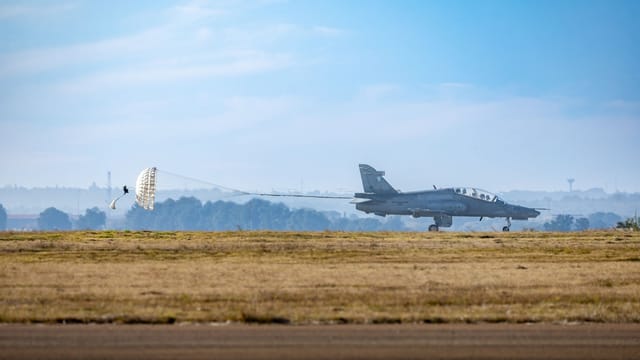 A military jet lands with a parachute deployed on a clear day at the airstrip.