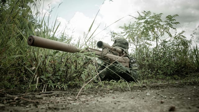 A camouflaged sniper with rifle hidden in grasslands during daytime surveillance.