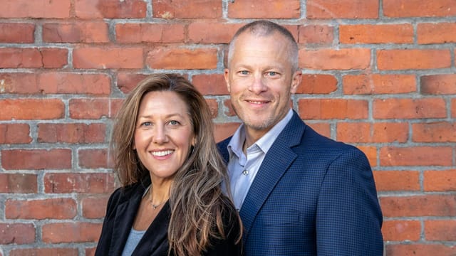 A cheerful middle-aged man and woman in business attire posing against a brick wall, depicting professionalism and partnership.