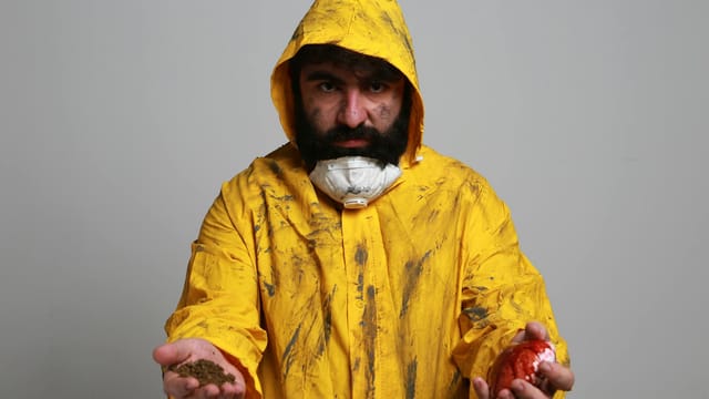 Portrait of a man in a yellow hazmat suit holding soil and a red apple, symbolizing contamination.