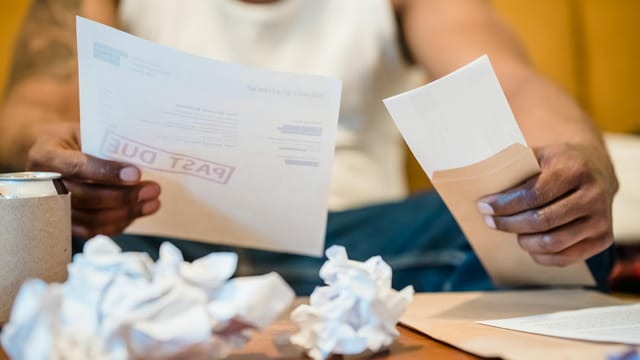 Man sitting indoors reviewing past due bills with crumpled papers on a coffee table.