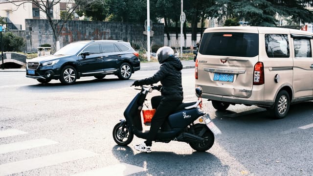 A person on a scooter navigates city traffic alongside cars in a busy urban street.
