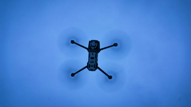 A drone seen flying in a clear blue sky, captured from below with motion blur on the rotors.