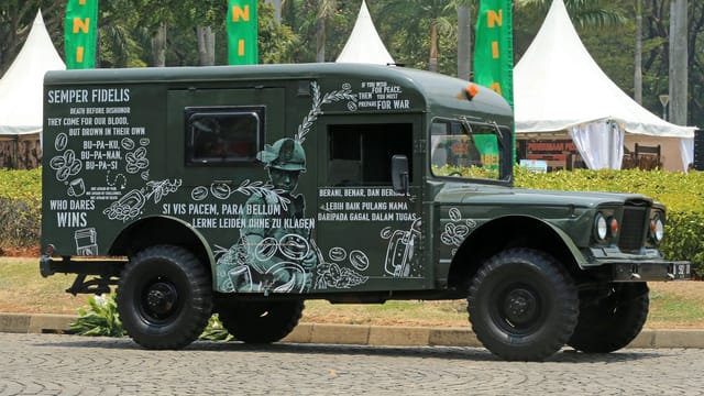 Green military vehicle with text and graphic decoration parked outdoors in an urban setting.