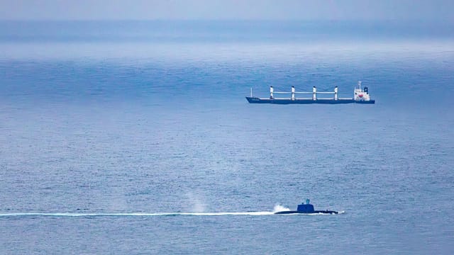 A cargo ship and submarine navigate the vast, open ocean under a clear sky.