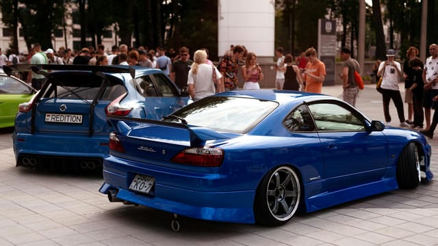 Rear view of two modified blue Nissan cars at an urban car show with a crowd gathered.