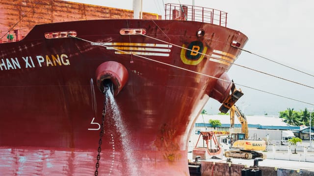 Front view of a large red cargo ship docked, releasing water, with construction equipment nearby.