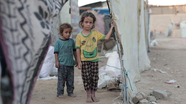 Two children stand together amidst a refugee camp setting in Gaza, showcasing resilience and hardship.