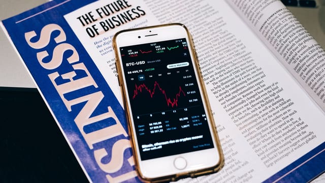 Close-up of a smartphone with a cryptocurrency graph placed on an open business magazine, highlighting economic trends.