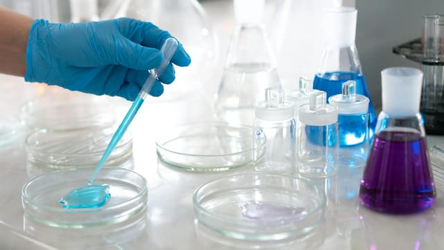 Close-up of a scientist's hand in glove conducting an experiment with petri dishes and colorful liquids in a lab.
