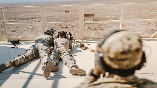 Soldiers in camouflage uniforms practicing sniper skills in a desert setting.