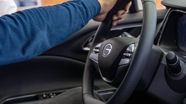 A person steers a car, focusing on a hand holding the steering wheel inside a vehicle.
