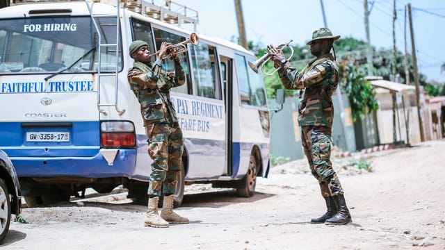 Two military personnel play trumpets beside a parked bus on a village dirt road during the day.