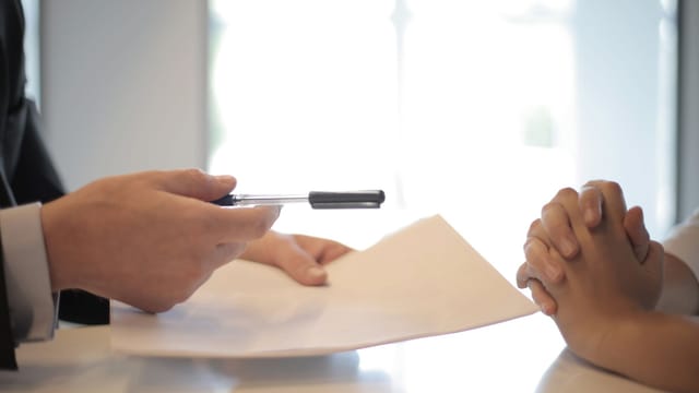 Side view of faceless formal man giving pen and paper to focused female with clenched hands at table on meeting