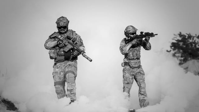 Black and white image of soldiers in a tactical operation amidst smoke and sand.