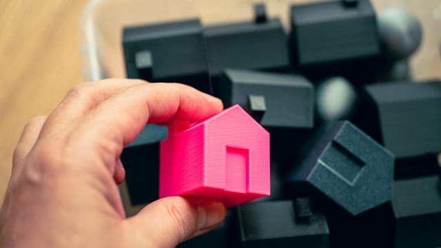 Hand selecting a pink house model from a box of miniature homes, symbolizing real estate choices.