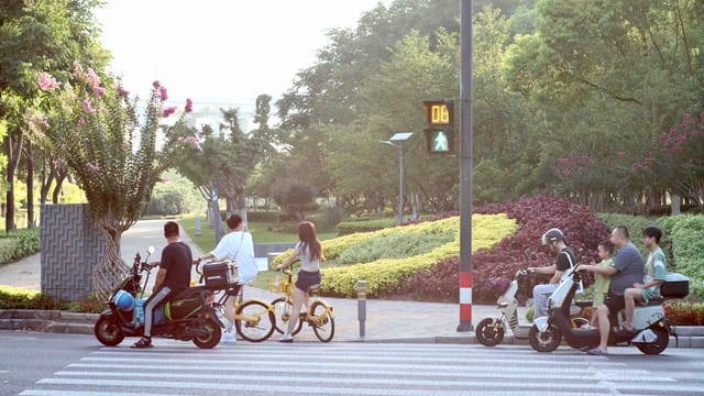 Group of adults and children on bicycles and motorbikes waiting at a crosswalk on a sunny day.