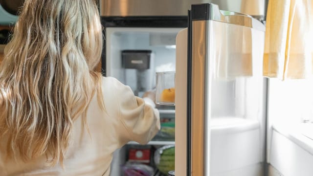 Back view of a blonde woman retrieving food from a refrigerator in a bright kitchen.