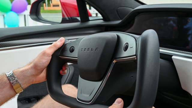 Hands gripping Tesla's minimalist yoke steering wheel in an electric vehicle interior.