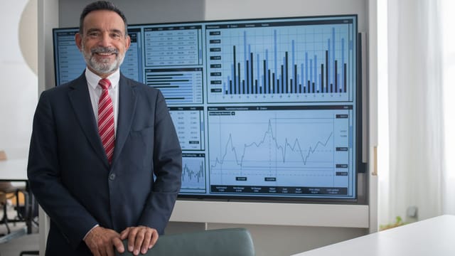 Smiling businessman in office analyzing financial graphs on a large screen, demonstrating professional success.